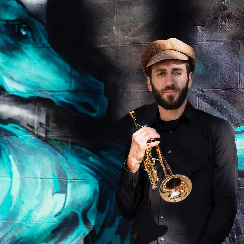 Ritratto di Alessandro Bottacchiari con tromba e cappello davanti a un murale blu
